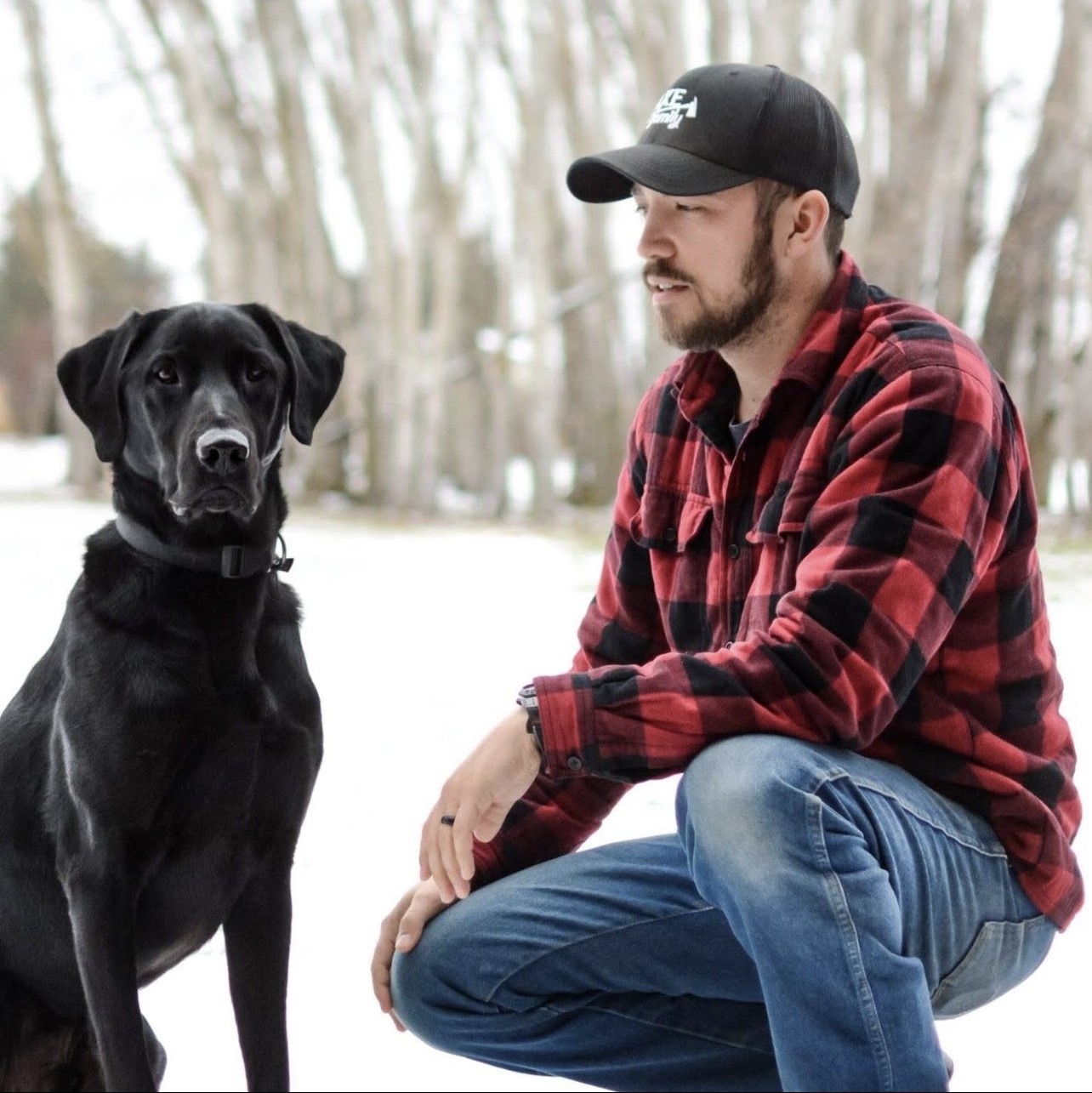 Cody kneeling in the snow next to his black Lab, both looking at the camera